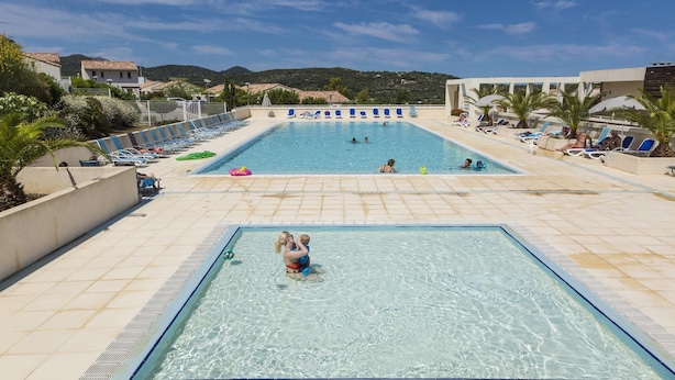 Ein Schwimmbecken mit Liegestühlen und Sonnenschirmen, umgeben von Gebäuden und Hügeln.