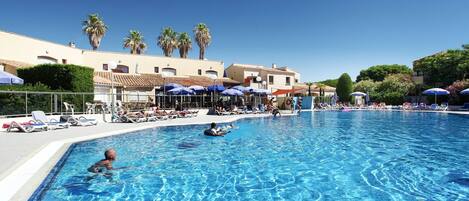 A swimming pool with lounge chairs and umbrellas, surrounded by buildings with palm trees.