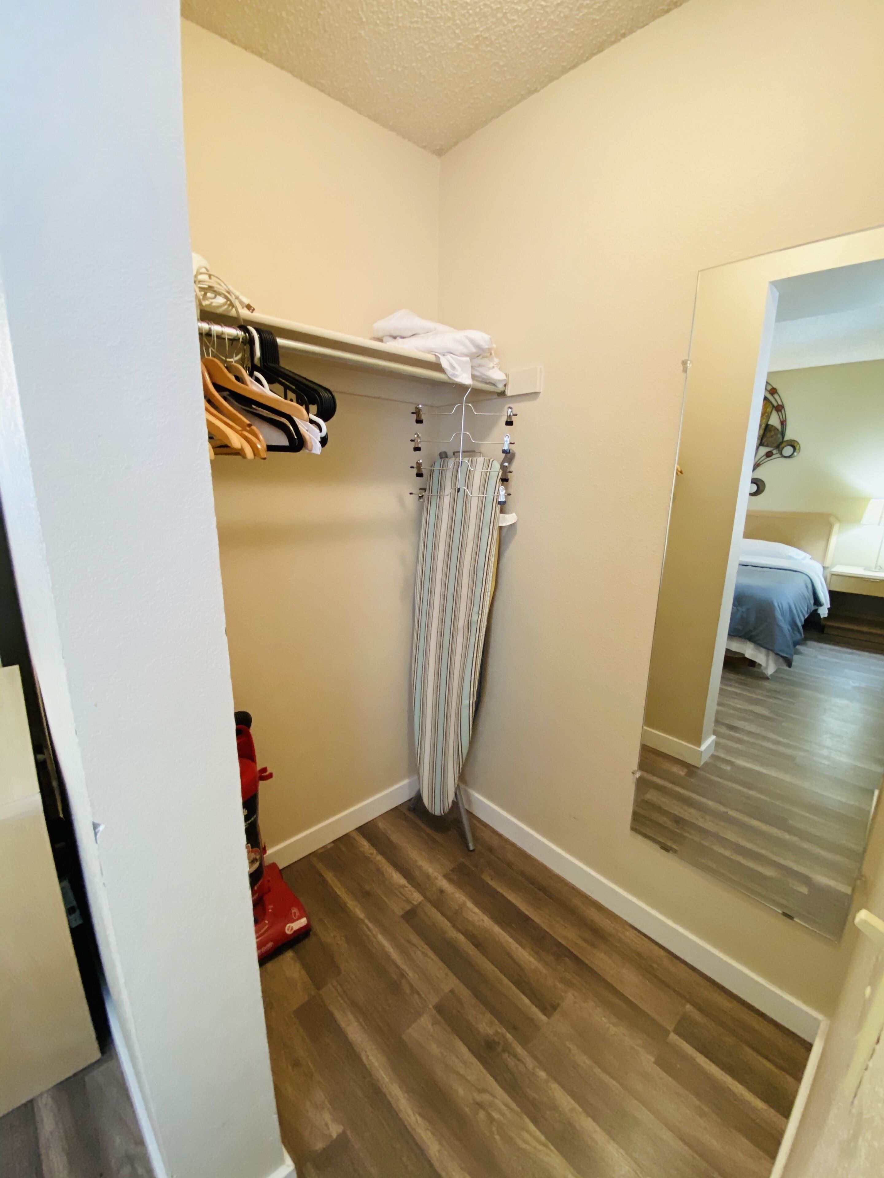 A bedroom with a wooden floor, a bed with a blue bedspread, a white closet with hanging clothes, and a striped curtain.
