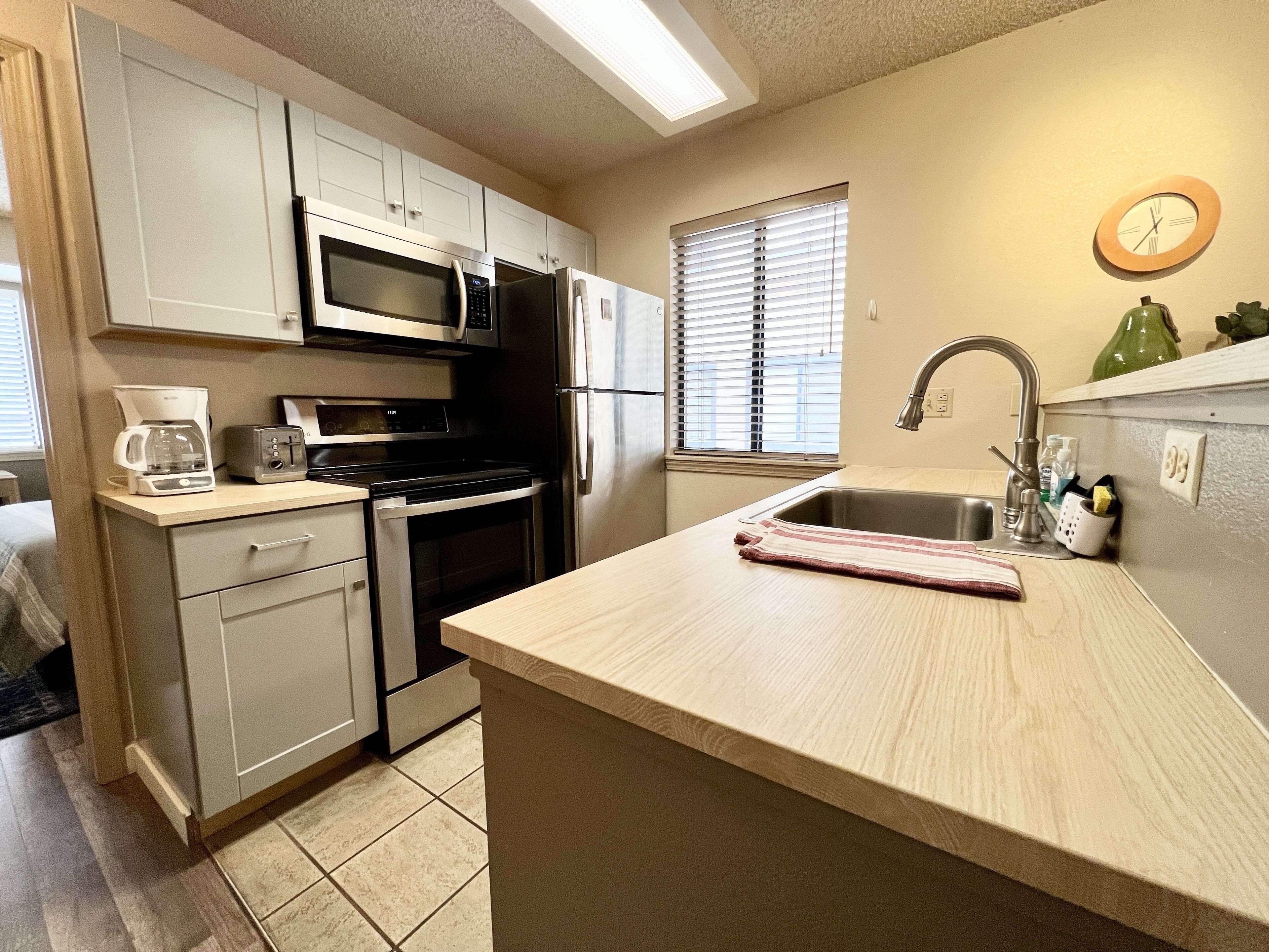 A compact kitchen with a microwave, oven, and refrigerator. There is a window with blinds, a sink with a faucet, and a clock on the wall.