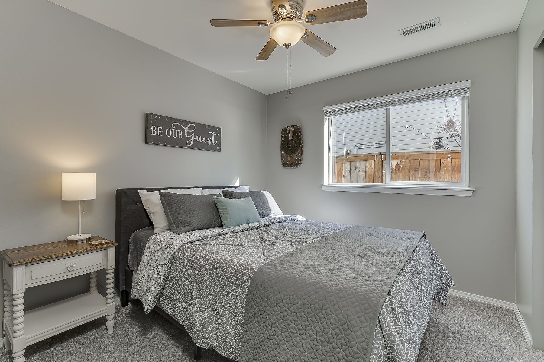 A bedroom with a bed, a nightstand, a wall-mounted sign, a window with a view, and a ceiling fan.