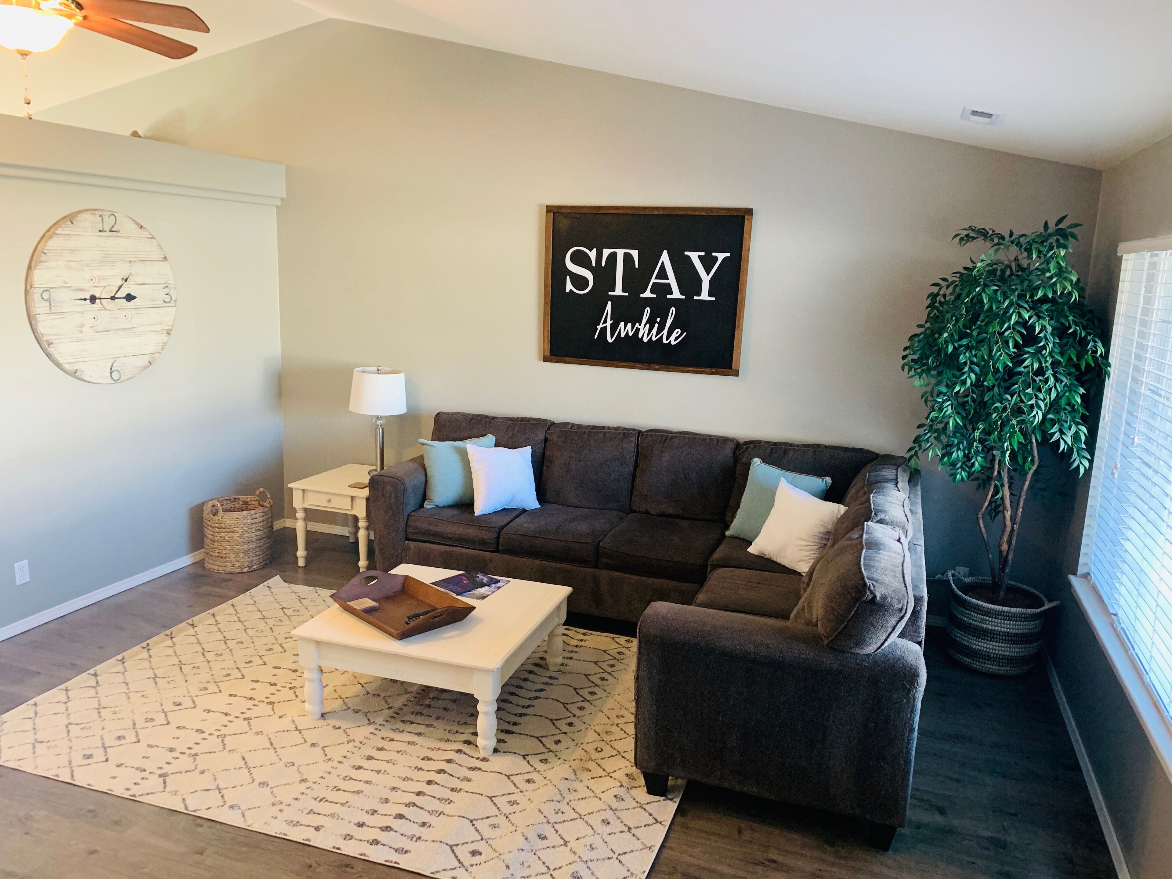 A cozy living room with a brown sofa, a white coffee table, and a large wall clock. A 'STAY AWAY' sign is displayed above the sofa.