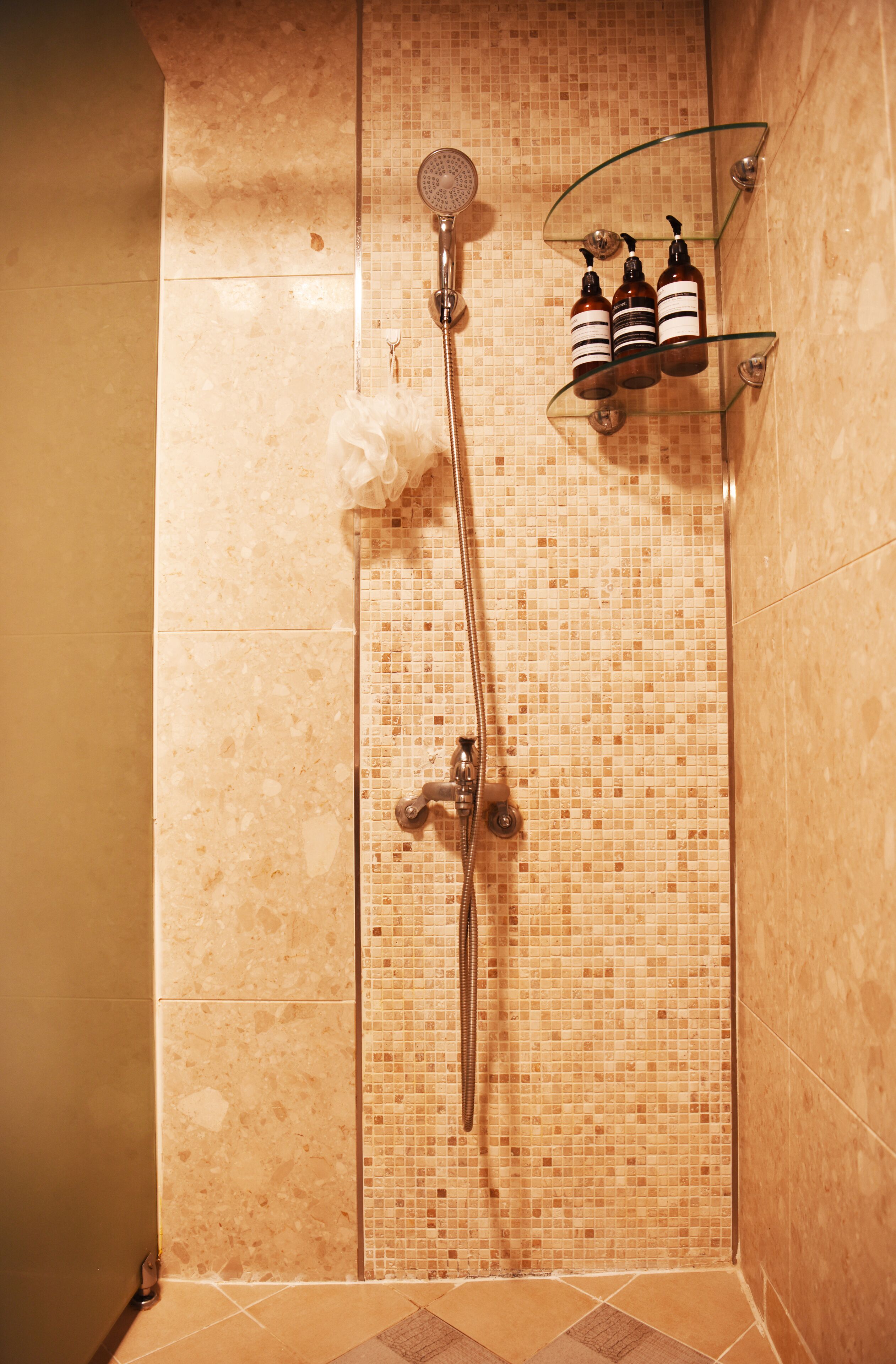 A shower with a tiled wall, a glass shelf with toiletries, and a showerhead.