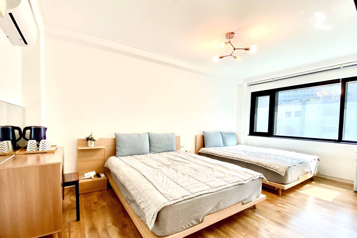 Un dormitorio moderno con dos camas, un aparador de madera, una cafetera y una ventana con vista a edificios.