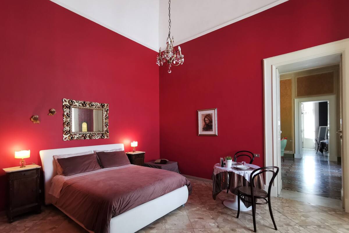 A bedroom with a bed, bedside tables, a chandelier, a mirror, a framed picture, and a dining area with a table and chairs.