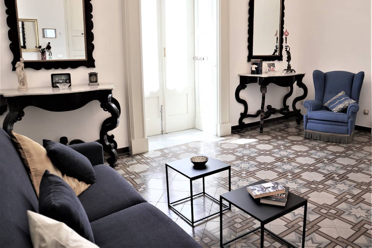 A living room with a sofa, armchairs, a coffee table, and two side tables. There are two large mirrors on the walls, a door leading to another room, and a patterned floor.