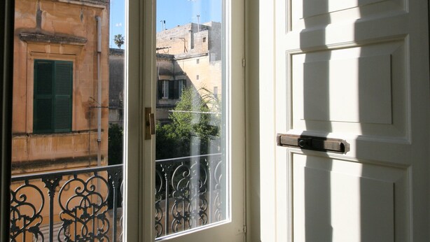 A view through a window with a balcony, featuring a building with a green shutter and a white door. - Chez Moi Lecce Charme B&B (Lecce)