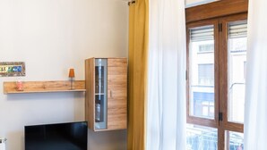 Un salon moderne avec un téléviseur à écran plat posé sur un meuble en bois, un meuble vitré, une étagère en bois avec une lampe et une grande fenêtre avec des rideaux.