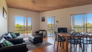 A room with a wooden ceiling, a sofa with patterned cushions, a dining table with chairs, and large windows with a view of greenery.