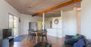 A spacious living area with a wooden ceiling, a dining table with chairs, a black leather sofa, and a television.