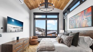 A bedroom with a large bed, a wooden dresser, a leather chair, and a large window with a view of mountains. - Ski-in/Ski-Out Luxury Schweitzer•Hot Tub•4 King Suites•Walk to Village•Ski Room (Sandpoint)