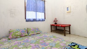 A room with a bed, a small table, a window with a curtain, and a wall-mounted light fixture. - JIO Homestay (Yogyakarta)