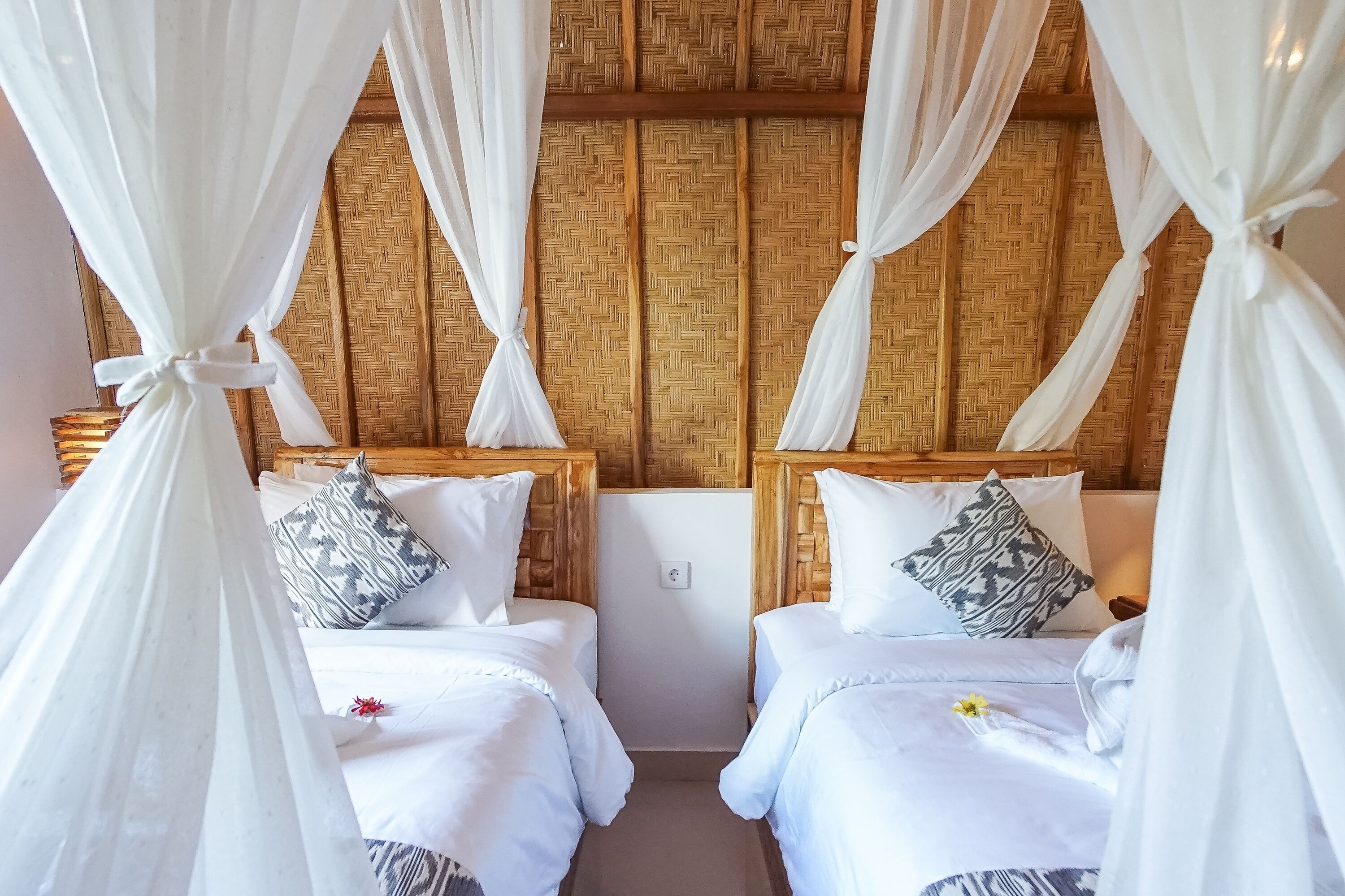 A room with two beds, white bedding, patterned pillows, and a woven bamboo headboard.
