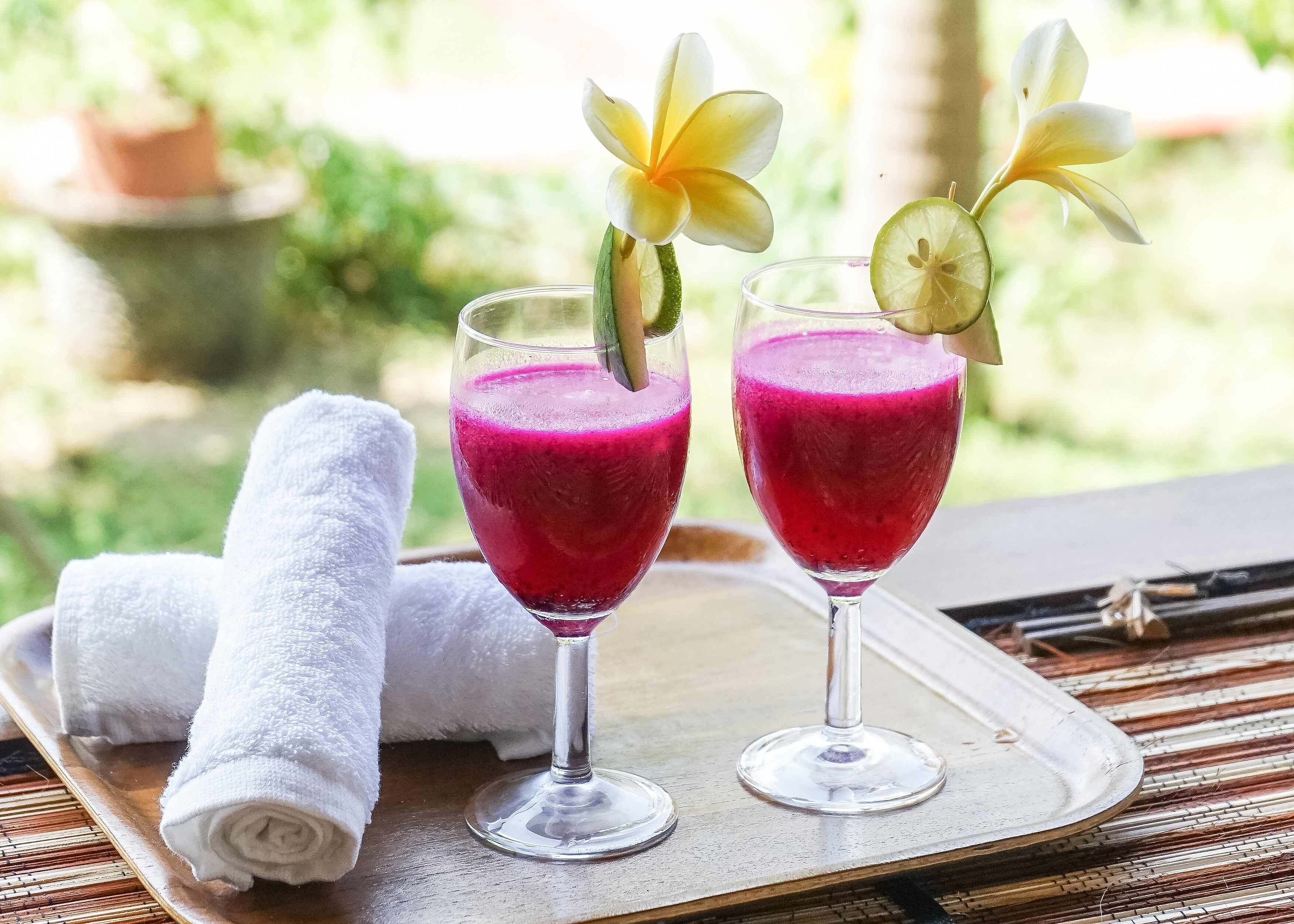 Two glasses of pink smoothie on a tray with a white towel and a flower.