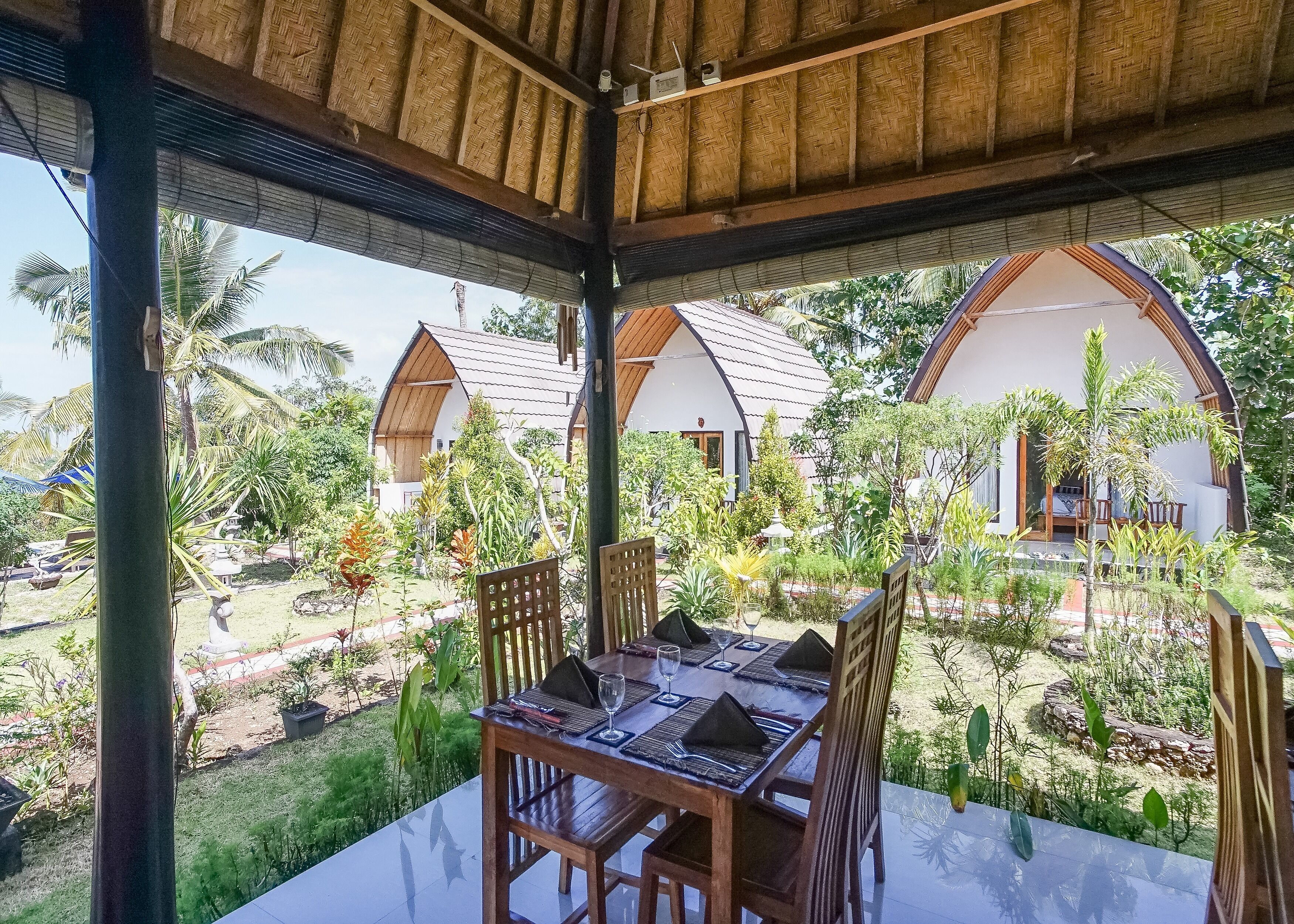 A covered outdoor dining area with a table set for two, surrounded by lush tropical vegetation and traditional-style buildings in the background.