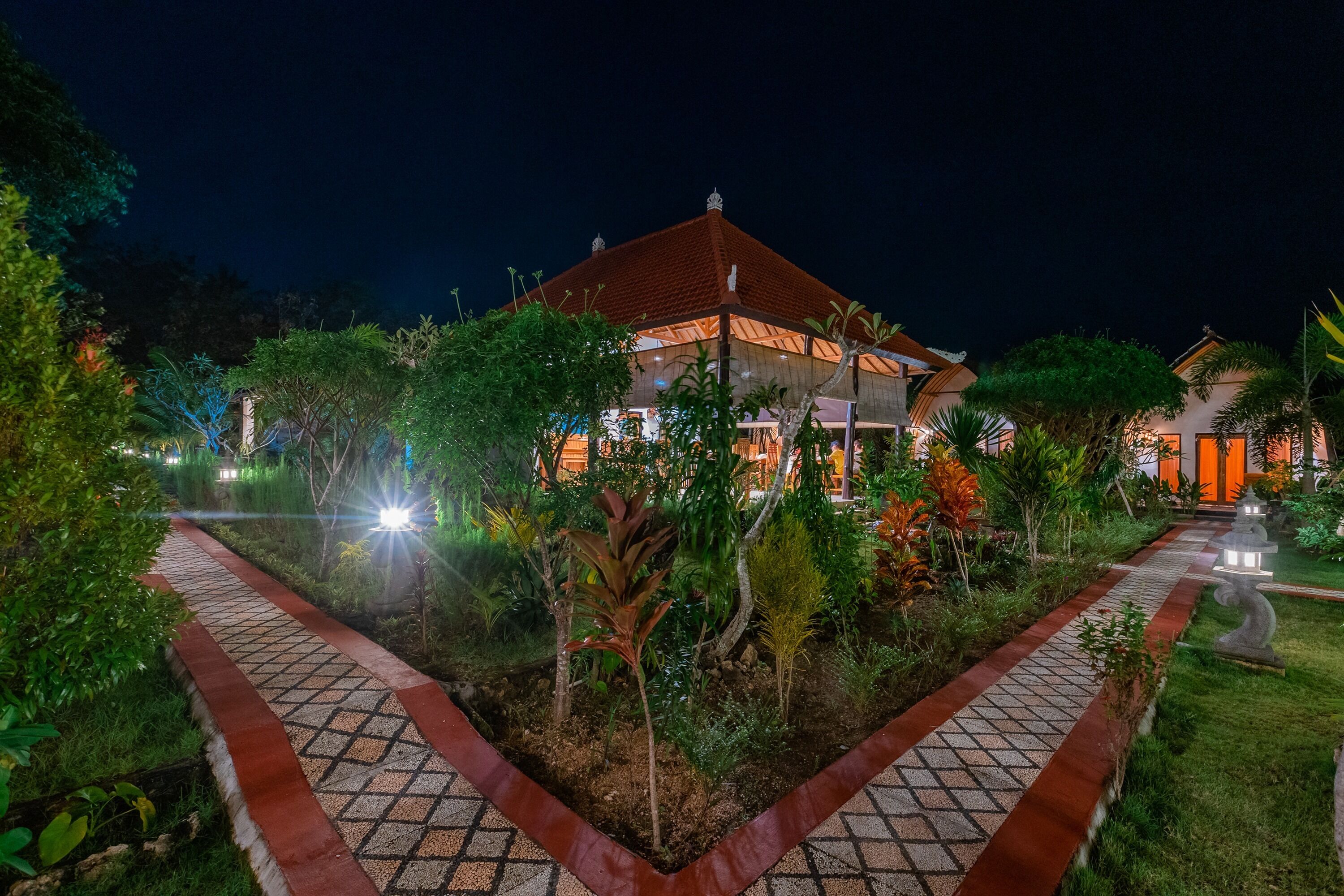 A well-lit building with a tiled pathway leading to it, surrounded by lush greenery.