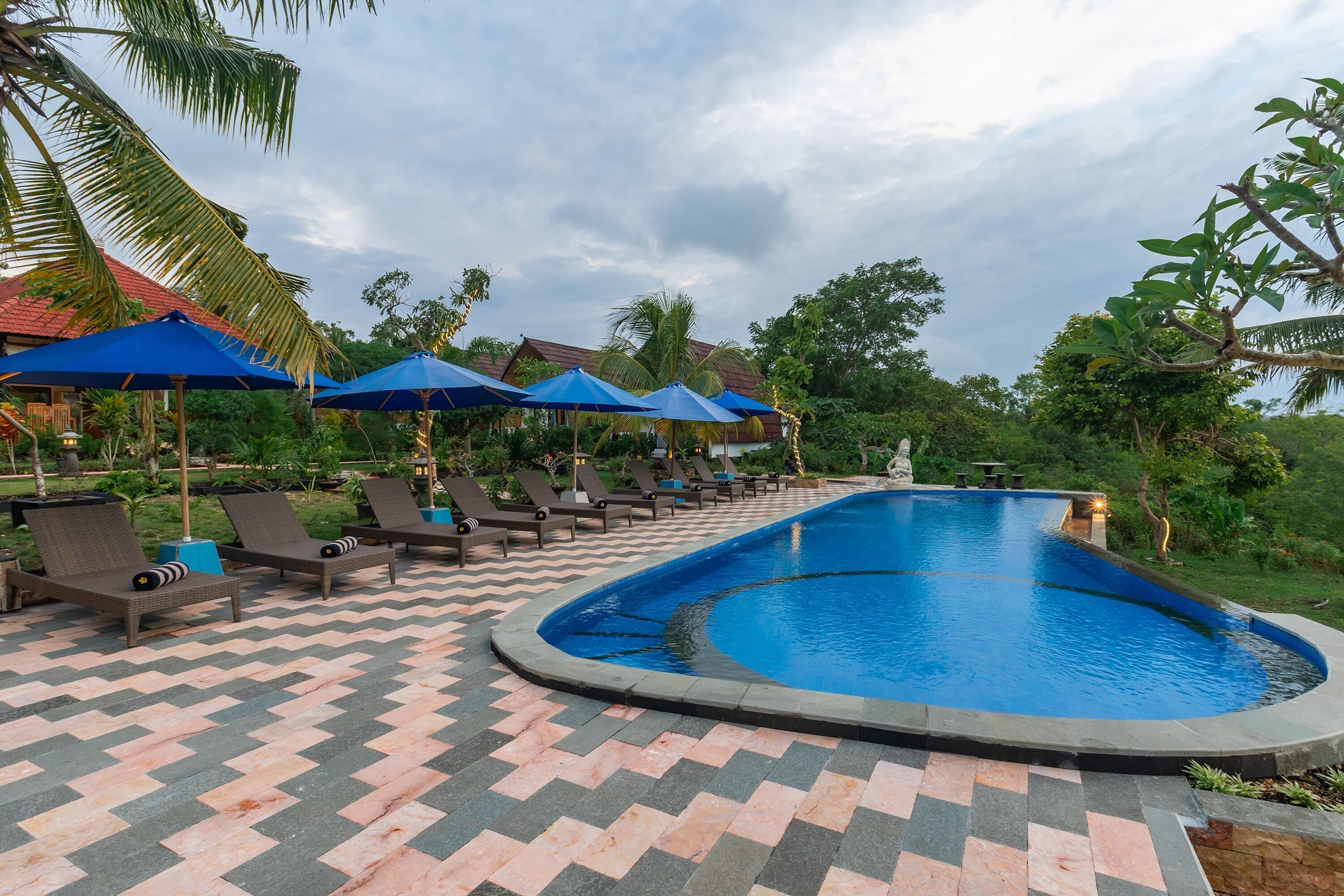 A swimming pool with lounge chairs and blue umbrellas, surrounded by palm trees and a herringbone-patterned walkway.