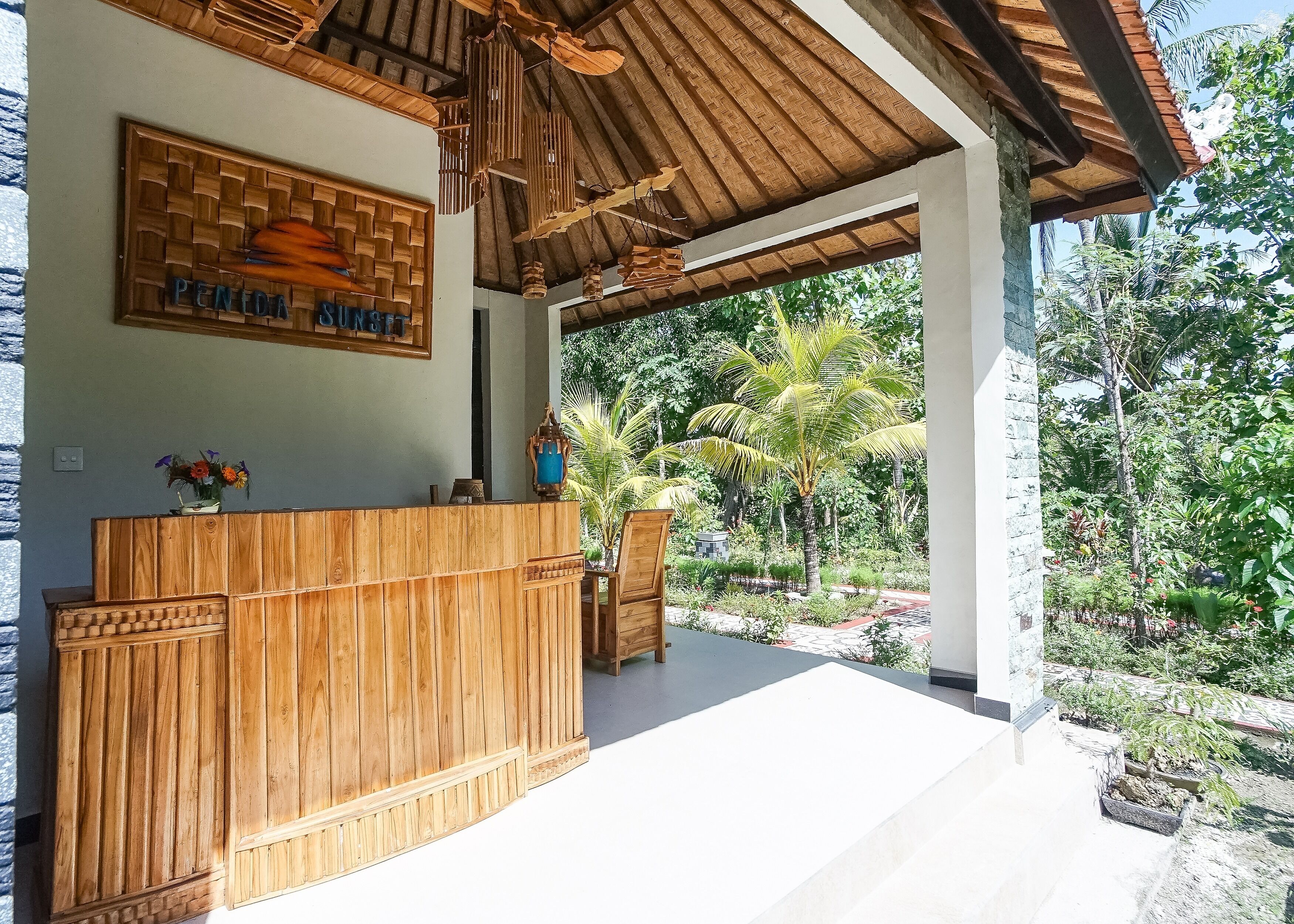 A reception area with a wooden reception desk, a sign above it, and a view of a lush garden with palm trees and various plants.