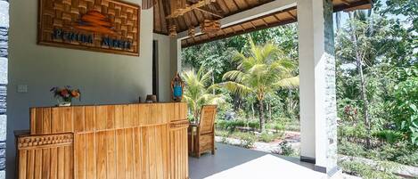 A reception area with a wooden reception desk, a sign above it, and a view of a lush garden with palm trees and various plants.