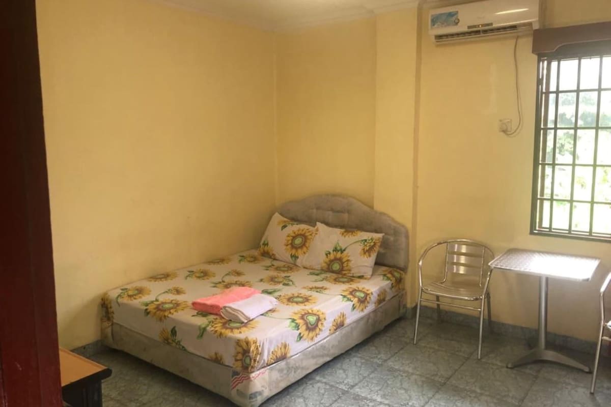A small, single-bed room with a floral bedspread, a chair, a desk, and an air conditioning unit.