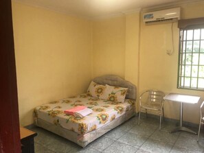 A small room with a bed, a chair, a desk, and an air conditioning unit. - Hotel Pelita Terang (Batam)