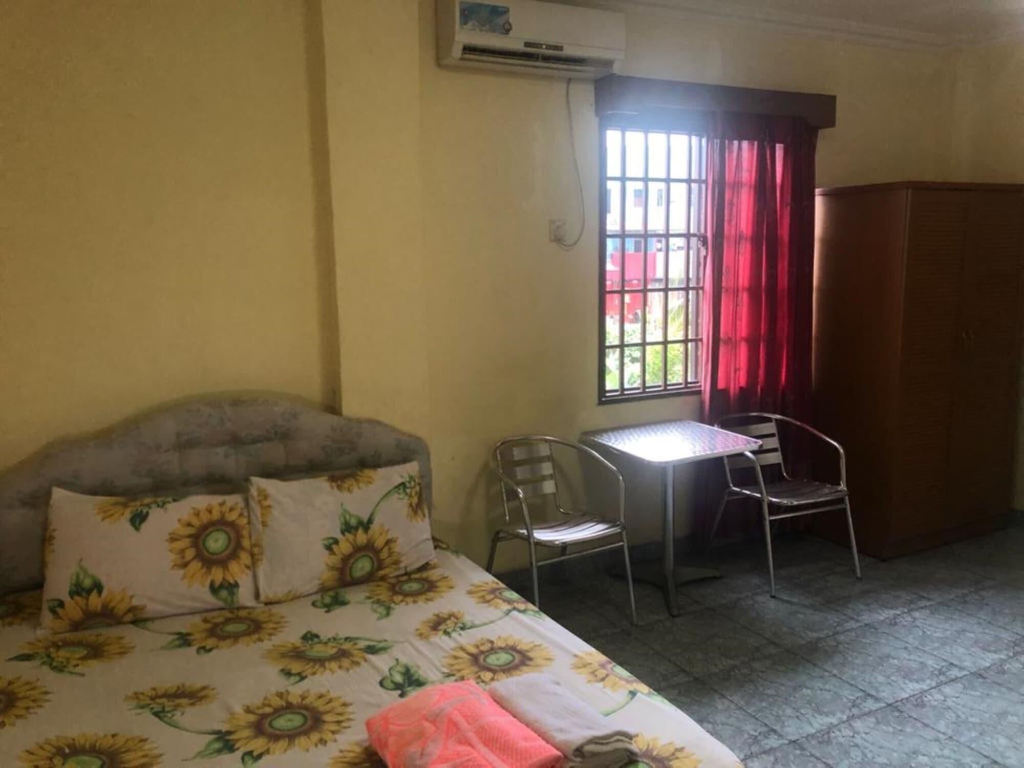 A room with a bed, two chairs, a small table, a window with curtains, and an air conditioning unit.