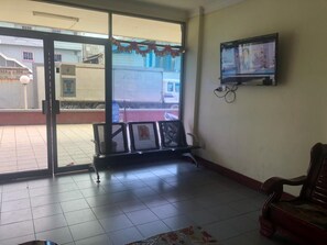 A room with a tiled floor, a bench, a television mounted on the wall, and a view through a glass door. - Hotel Pelita Terang (Batam)