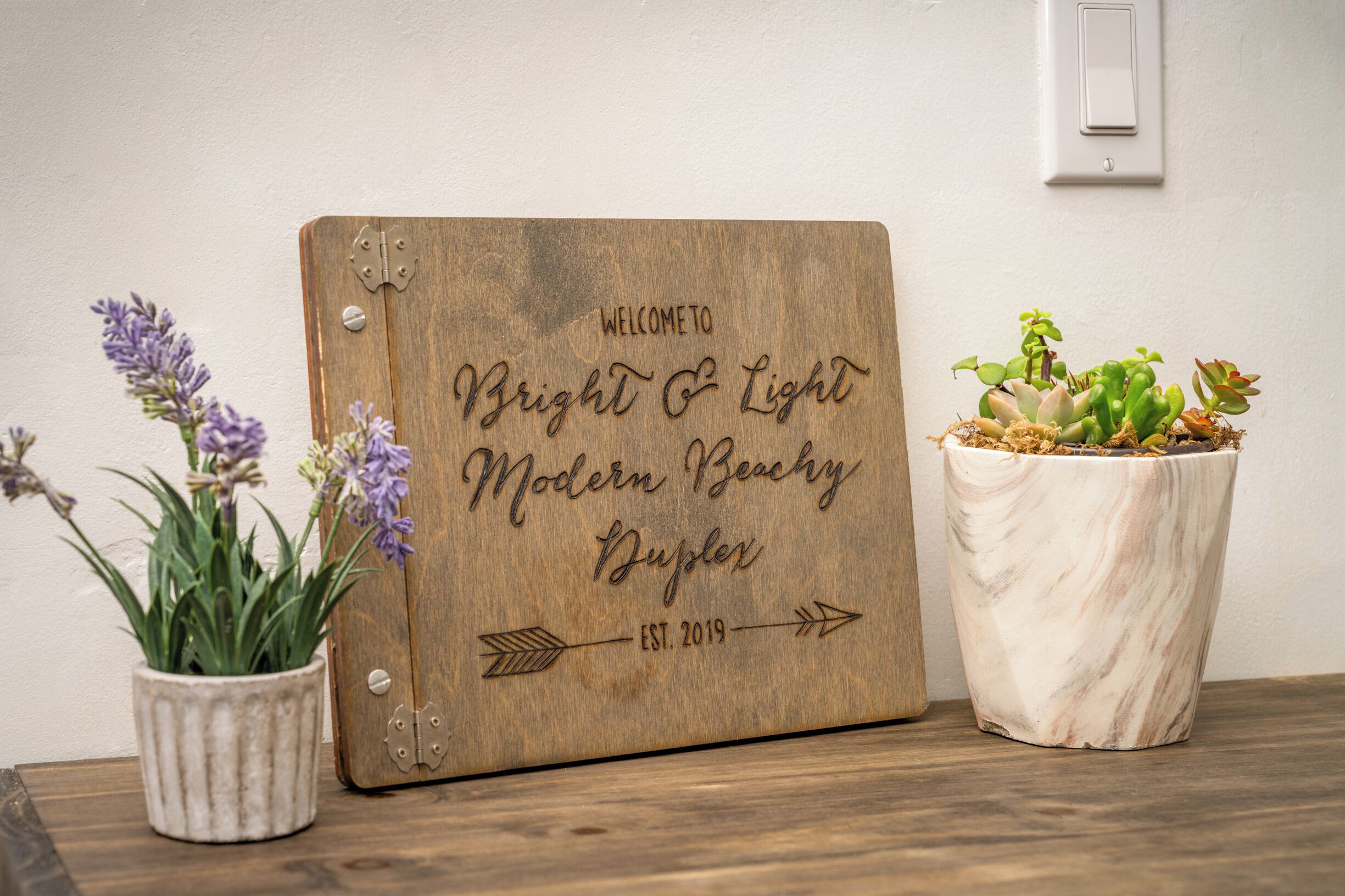 A wooden sign with "Bright & Light Modern Beachy Duplex" and "EST. 2019" on a wall.