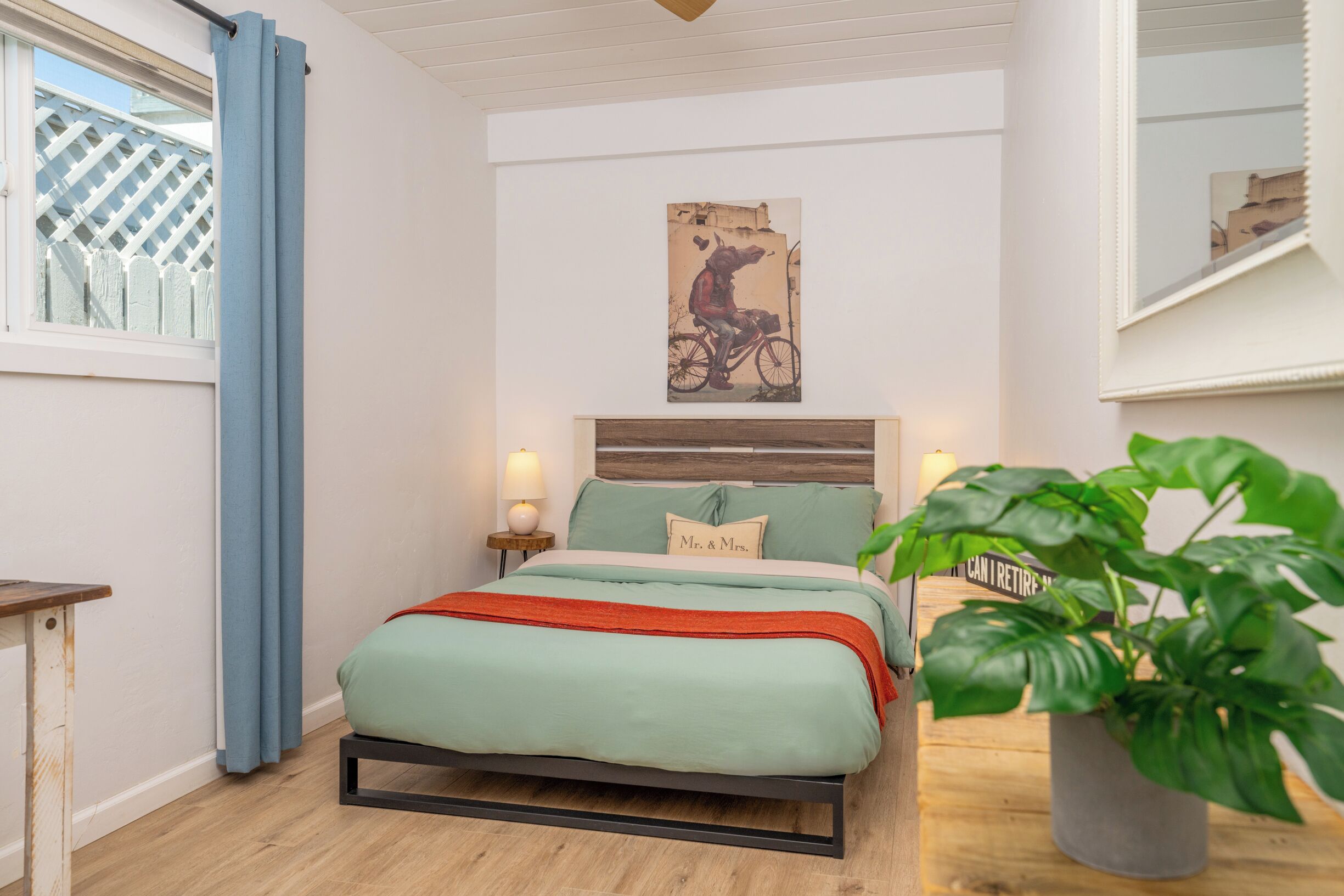 A bedroom with a bed, a headboard, a nightstand, a lamp, a potted plant, a window with curtains, and a wall-mounted picture.