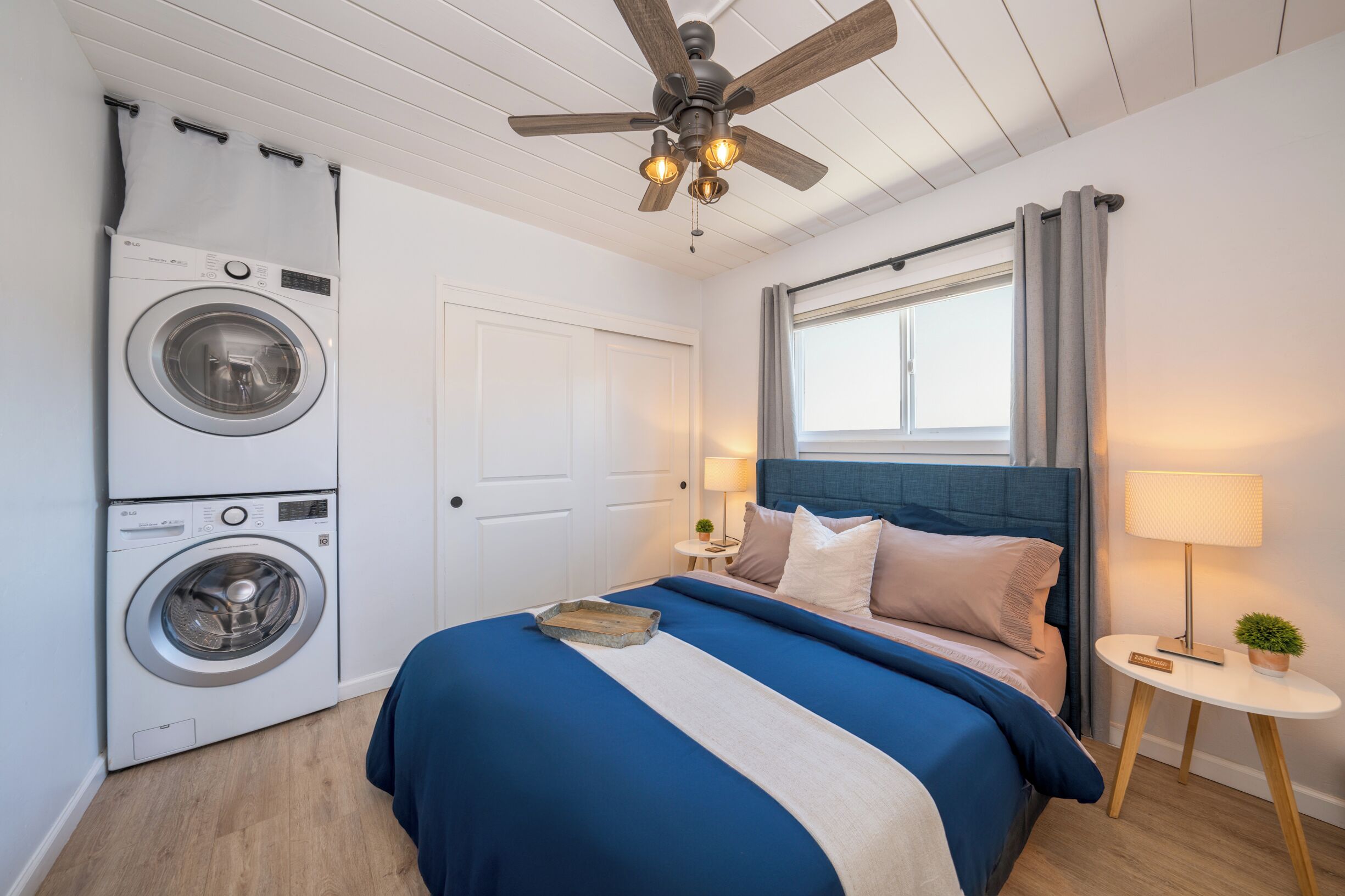 A bedroom with a washing machine, a bed with blue bedding, a ceiling fan, and a window with curtains.