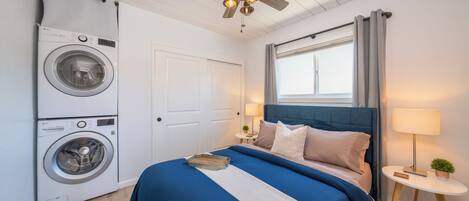 A bedroom with a washing machine, a bed with blue bedding, a ceiling fan, and a window with curtains.