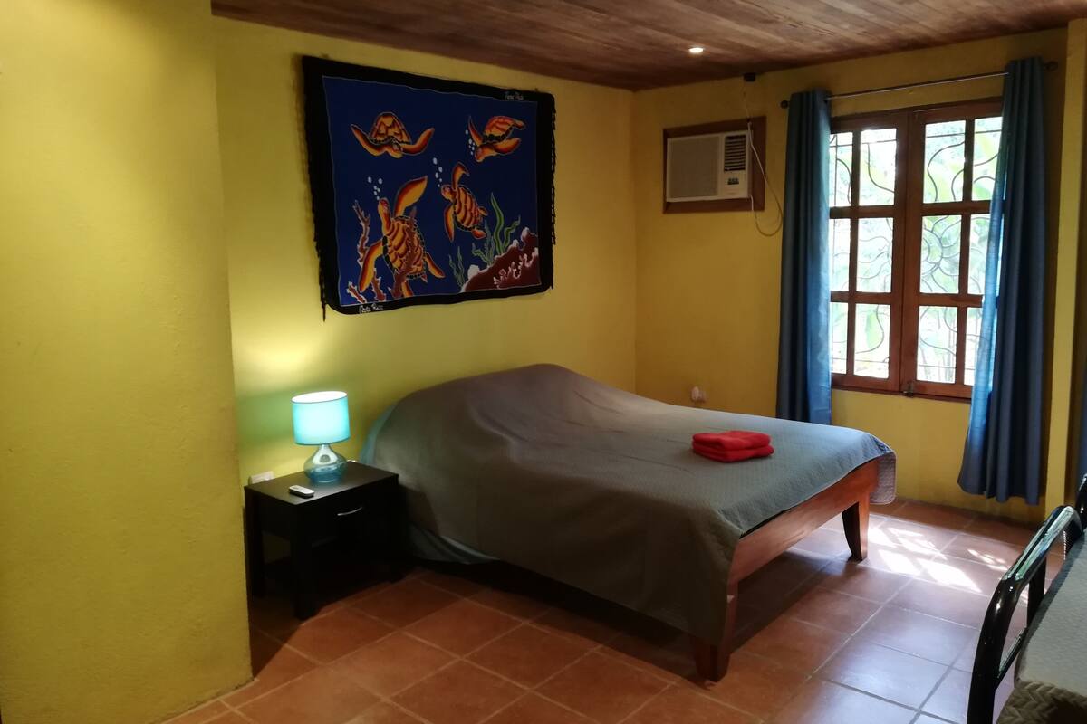 A bedroom with a bed, a nightstand, a wall-mounted air conditioner, and a window with curtains.