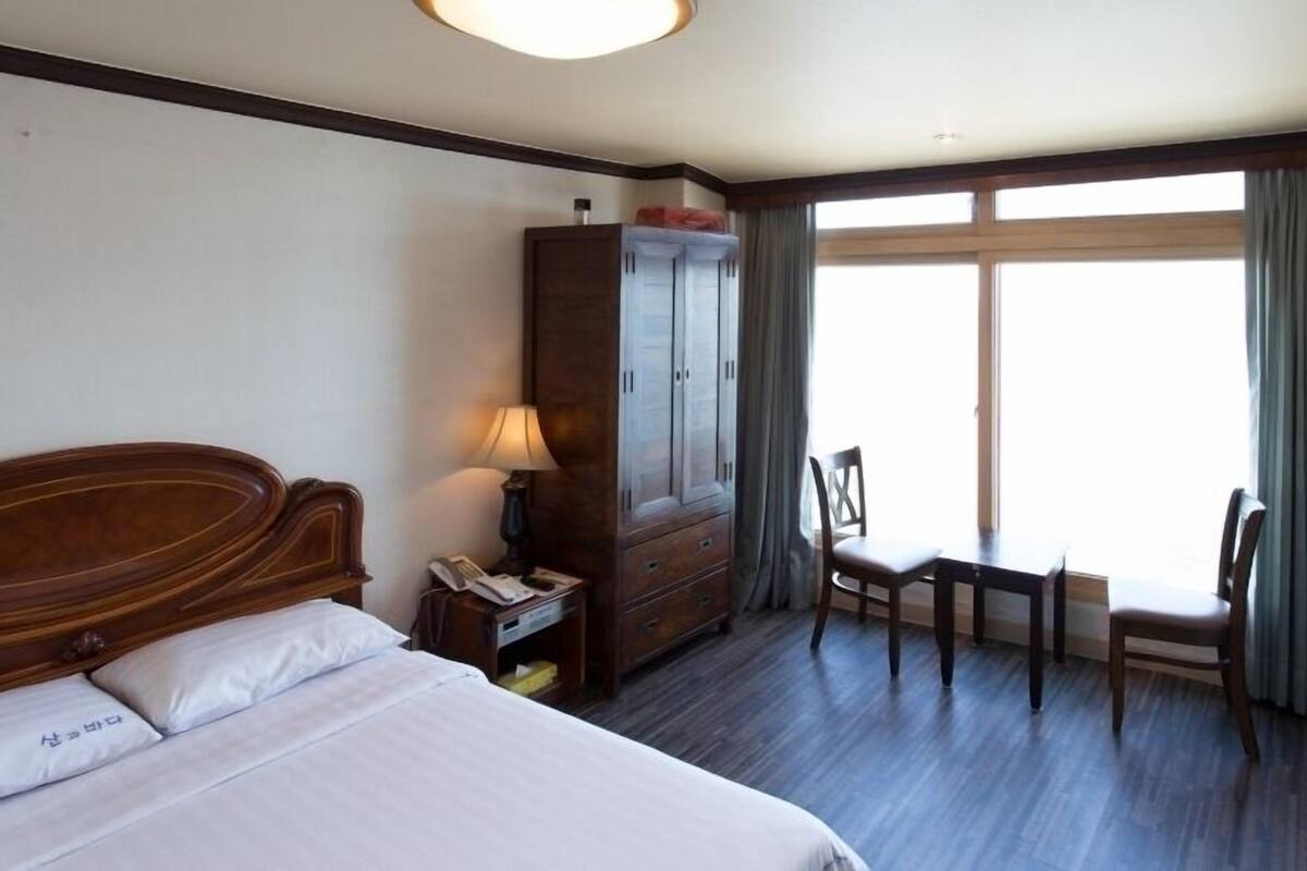 A hotel room with a wooden bed, a desk with a chair, a wardrobe, and a large window with curtains.
