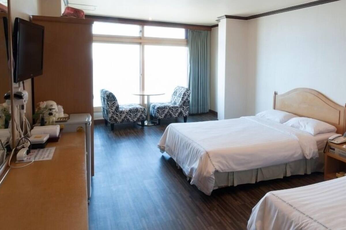 A hotel room with two beds, a desk with a telephone, a chair, a TV, and a window with curtains.