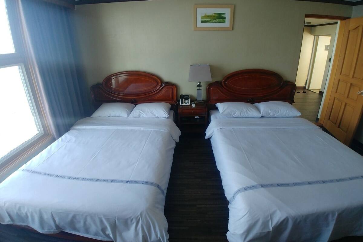 A hotel room with two single beds, a wooden headboard, a bedside table with a lamp, and a framed picture on the wall.