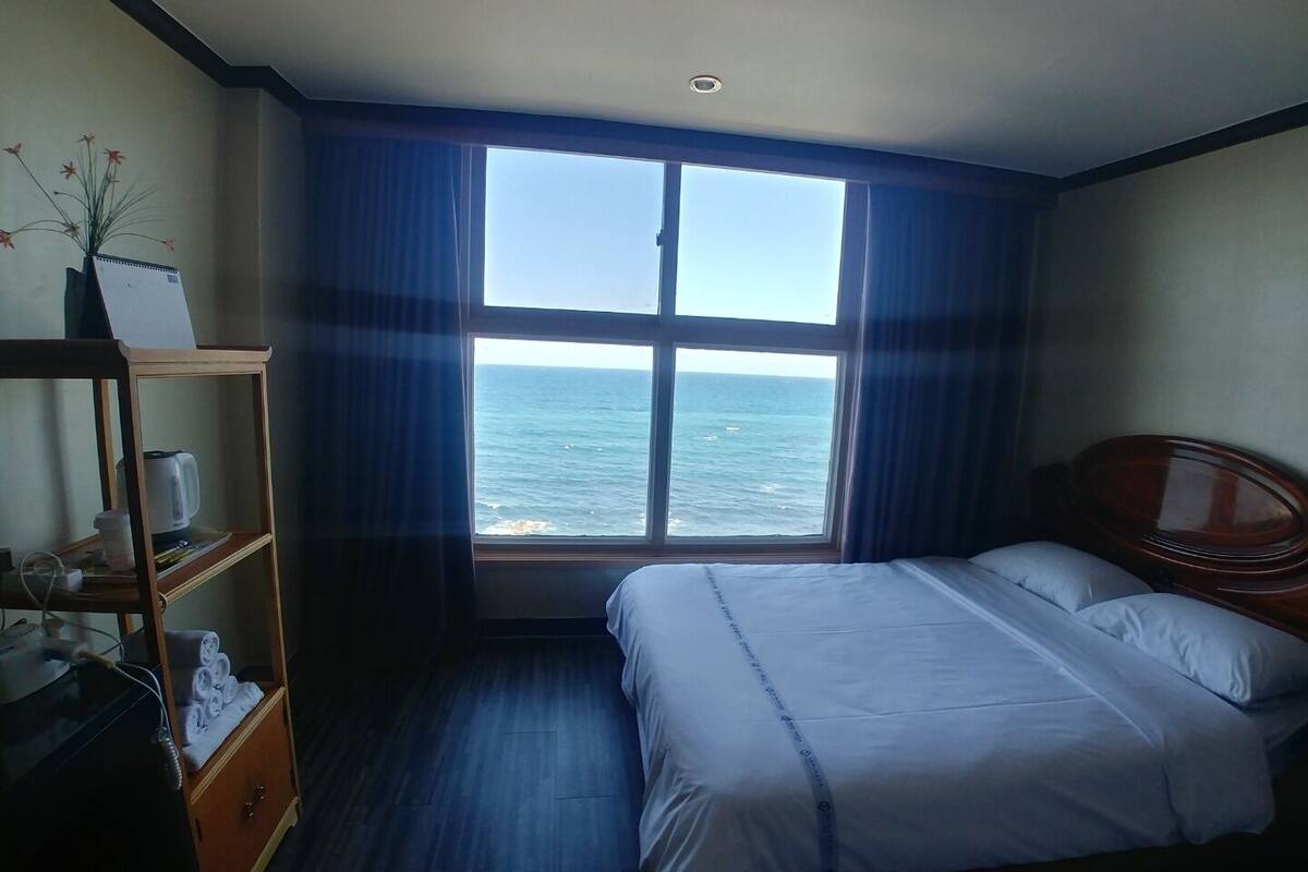 A hotel room with a bed, a wooden nightstand, a shelf with towels, and a window with a view of the ocean.