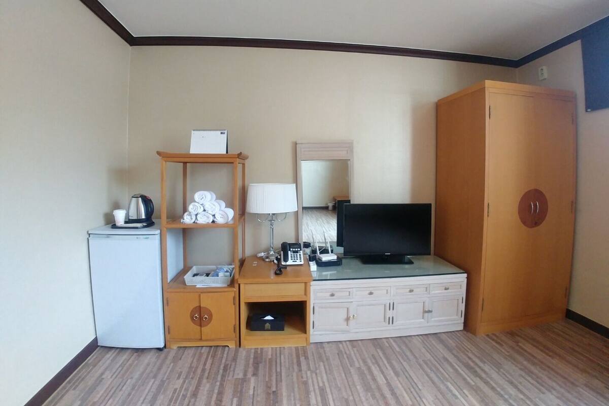 A hotel room with a bed, a TV, a desk with a lamp, a wardrobe, and a small refrigerator.