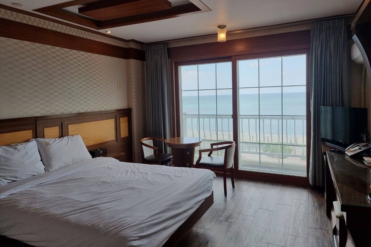 A hotel room with a large bed, a small table, a chair, a TV, and a balcony with a view of the sea.