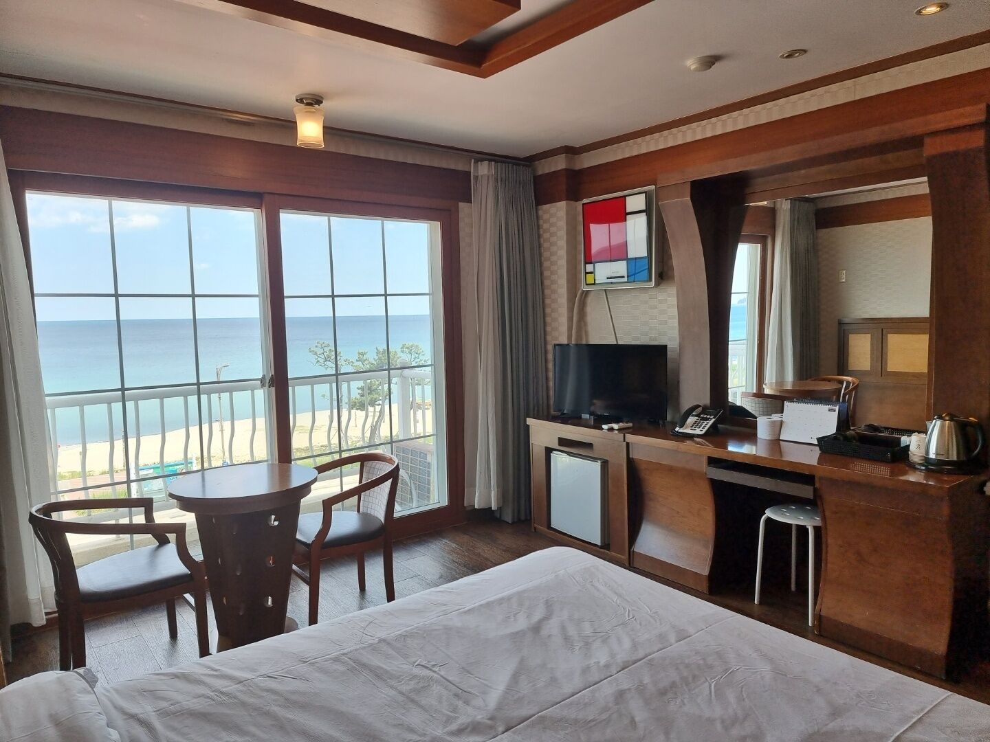 A hotel room with a bed, a desk with a TV, a phone, and a small table with chairs. There is a large window with a view of the beach and ocean.