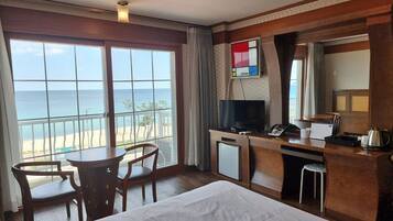 A hotel room with a bed, a desk with a TV, a phone, and a small table with chairs. There is a large window with a view of the beach and ocean.