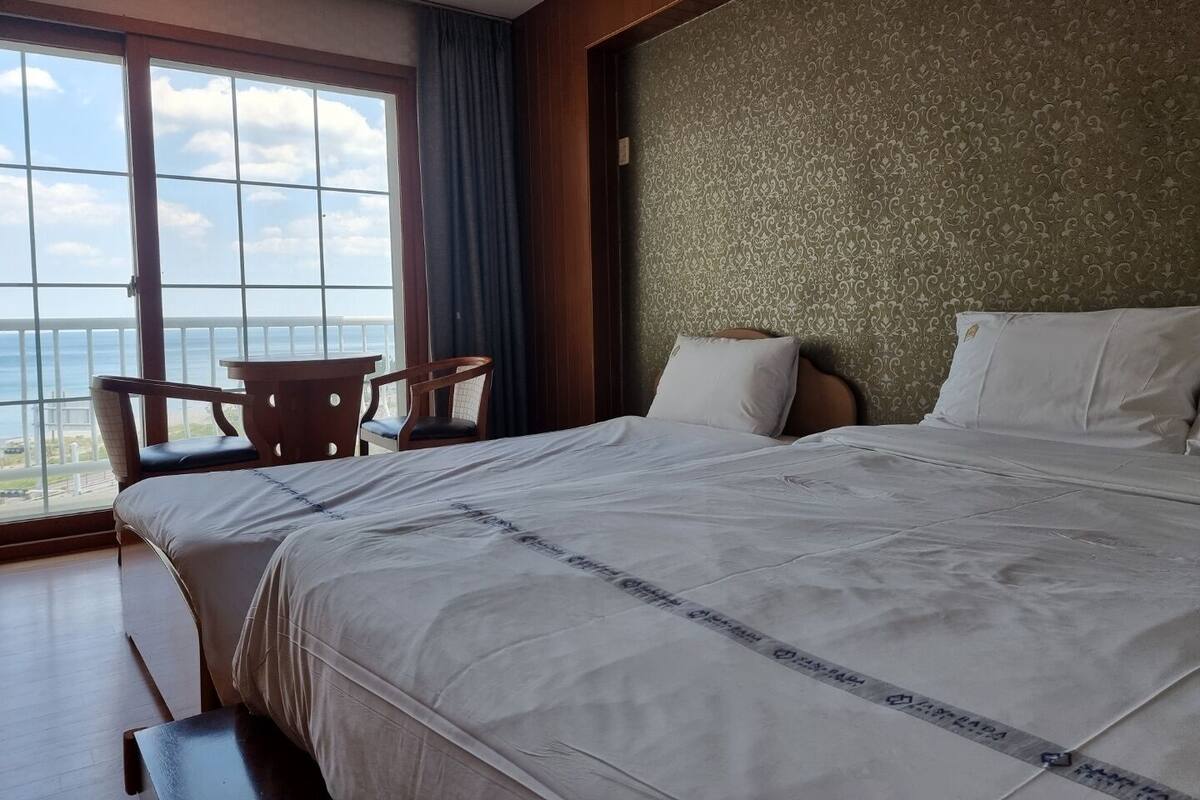 A hotel room with a large bed, a chair, a small table, and a view of the ocean through large windows.