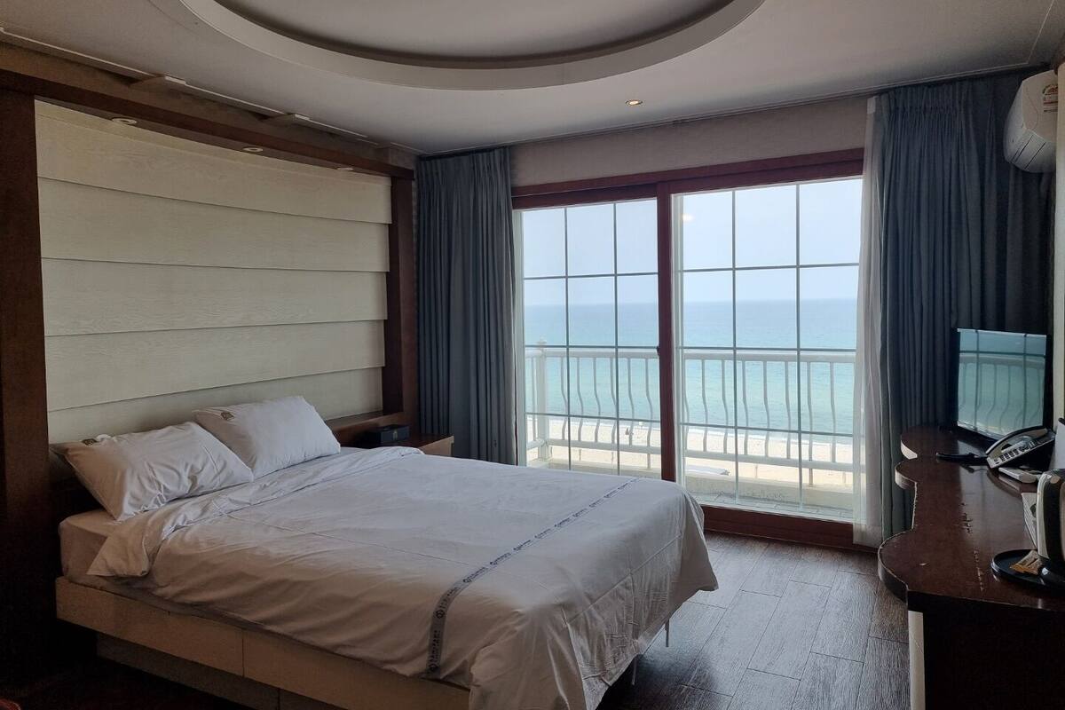 A hotel room with a large bed, a desk with a phone, and a view of the ocean through a large window.