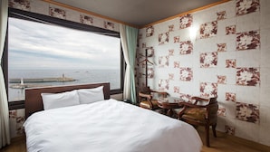 Une chambre d’hôtel avec un grand lit, un bureau et une chaise. La fenêtre offre une vue sur la mer et un phare.