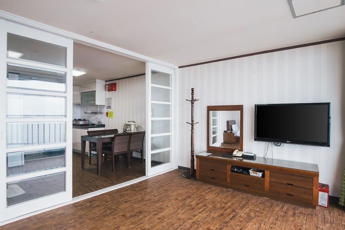 A room with a wooden floor, a TV mounted on the wall, a wooden cabinet, and a sliding glass door leading to a kitchen area.