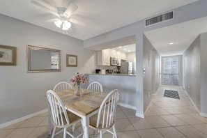 A dining area with a wooden table and white chairs, a ceiling fan, and a kitchen in the background. - 2/2 Ground Unit 10 Min to beach on golf course (Venice)