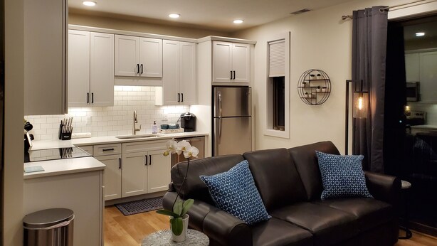 A modern kitchen with white cabinets, a stainless steel refrigerator, and a dining area with a black leather couch and patterned pillows. - Capri 107 Downtown Traverse City  (Traverse City)