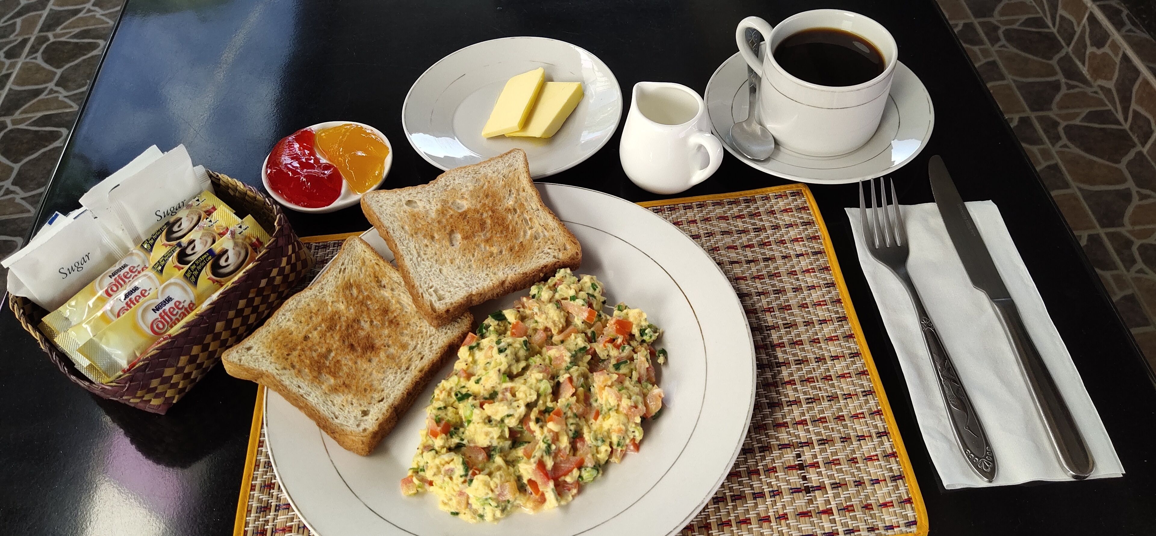 A breakfast setting with toast, scrambled eggs, butter, jam, coffee, and a basket of coffee packets.