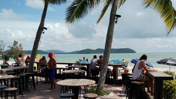 An outdoor dining area with wooden tables and chairs, overlooking a beach with umbrellas and people. - Ocean Lodge (Langkawi)