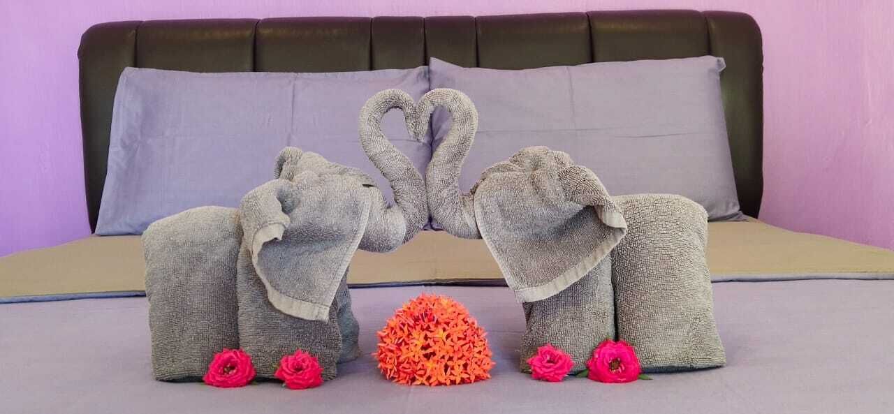 A bed with a grey blanket and two towels shaped like elephants with hearts, and a small orange flower arrangement.