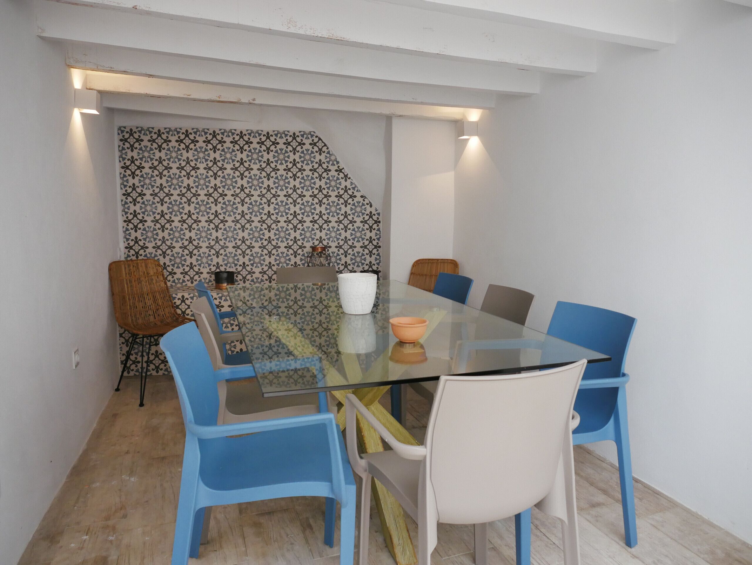 Un espace repas moderne avec une table en verre entourée de chaises bleues et blanches, un mur à motifs et un petit objet décoratif posé sur la table.